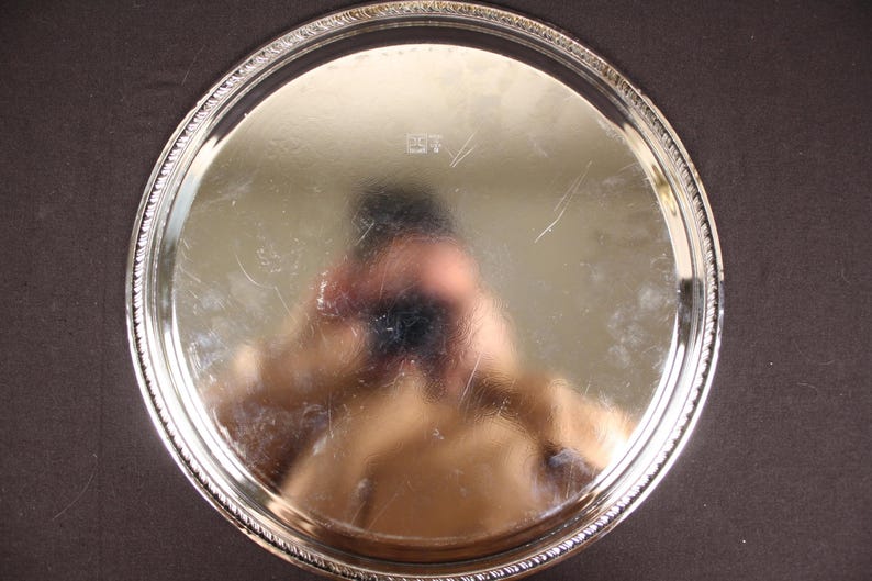 May include: A round, silver-colored serving tray with a decorative beaded edge. The tray's surface reflects light, showing a distorted reflection. The tray is set against a dark background, highlighting its bright finish.