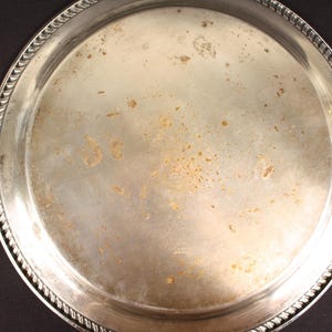 May include: A round, silver-colored metal tray with a decorative rope-like edge. The tray has visible signs of wear and tear, including discoloration and spots. The tray is set against a dark background.