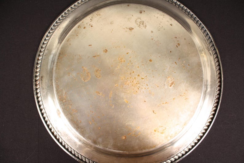 May include: A round, silver-colored metal tray with a decorative rope-like edge. The tray has visible signs of wear and tear, including discoloration and spots. The tray is set against a dark background.
