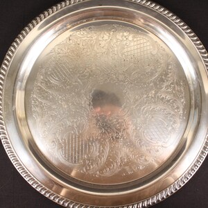 May include: A round, silver-colored serving tray with an ornate, engraved floral pattern. The tray has a decorative, rope-like edge. The reflective surface of the tray shows a distorted reflection of the surroundings.