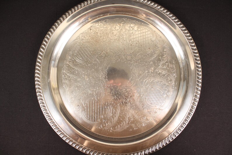 May include: A round, silver-colored serving tray with an ornate, engraved floral pattern. The tray has a decorative, rope-like edge. The reflective surface of the tray shows a distorted reflection of the surroundings.