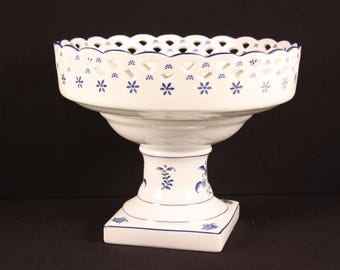 May include: A white ceramic pedestal bowl with blue floral accents. The bowl has a scalloped rim with cut-out designs and small blue flower patterns. The pedestal base is square and decorated with blue floral motifs. A decorative piece for serving or display.