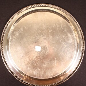 May include: A round, silver-colored serving tray with a decorative rope-like border. The tray has a smooth, reflective surface with minor scratches. The tray is likely made of metal and is suitable for serving food or drinks.