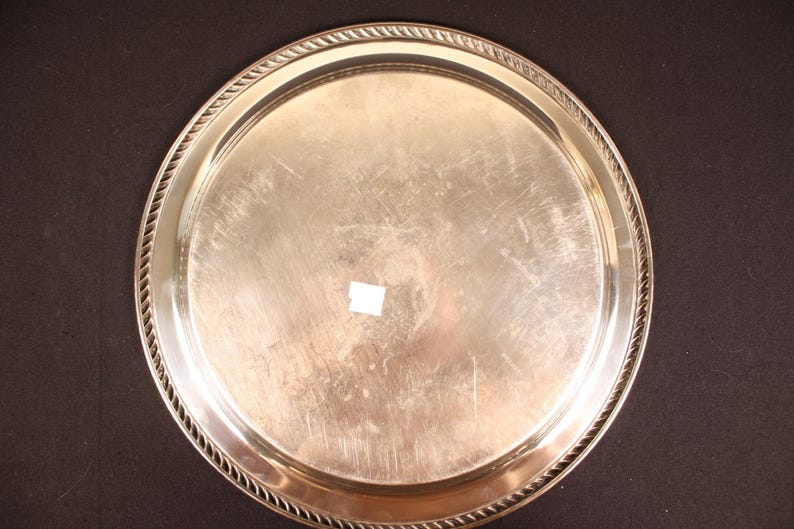 May include: A round, silver-colored serving tray with a decorative rope-like border. The tray has a smooth, reflective surface with minor scratches. The tray is likely made of metal and is suitable for serving food or drinks.