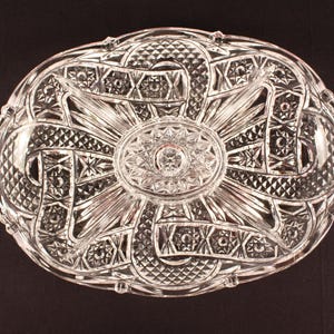 May include: An oval clear glass serving dish featuring an intricate pressed pattern of intersecting ribbons, diamond cross-hatching, and a central starburst motif. The dish has two integrated side handles and a scalloped, textured rim design.