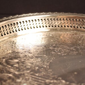May include: Close-up of a silver serving tray with ornate floral and scrollwork detailing. The tray's rim features a decorative pierced design. The reflective surface of the tray catches the light, highlighting the intricate patterns and textures.
