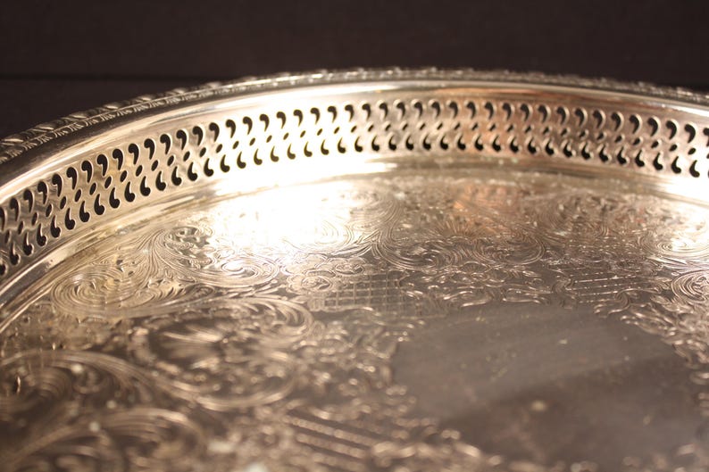 May include: Close-up of a silver serving tray with ornate floral and scrollwork detailing. The tray's rim features a decorative pierced design. The reflective surface of the tray catches the light, highlighting the intricate patterns and textures.