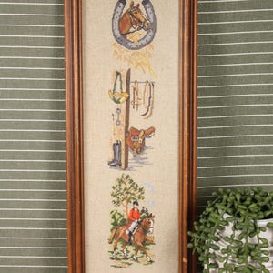 May include: A framed cross-stitch equestrian scene. The artwork features a horseshoe, riding equipment, and a rider on horseback. The frame is made of wood and the background is a neutral color.