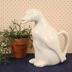 May include: A white ceramic duck-shaped pitcher with a handle, beak, and feet. The pitcher is glossy and sits on a blue surface. A terracotta pot with green plants is to the left of the pitcher.