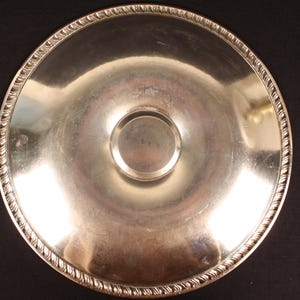 May include: A silver-colored, round serving tray with a decorative rope-like edge. The tray has a slightly concave center with a smaller, raised circular area. The reflective surface suggests it is made of metal.
