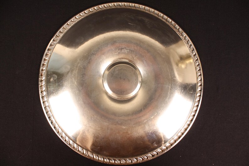 May include: A silver-colored, round serving tray with a decorative rope-like edge. The tray has a slightly concave center with a smaller, raised circular area. The reflective surface suggests it is made of metal.