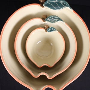 May include: Set of three apple-shaped ceramic bowls. The bowls are nested, with a cream-colored interior and a peach-colored rim. Each bowl has a brown stem and a green leaf detail, creating a charming, fruit-themed design.