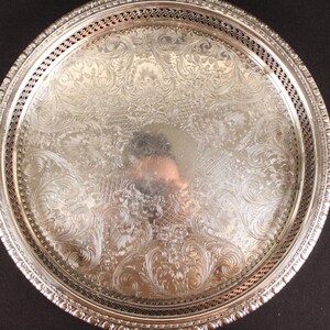 May include: A round, silver-colored serving tray with an ornate, embossed floral design. The tray has a decorative pierced border and a reflective surface. The tray is likely made of metal and is suitable for serving food or displaying items.
