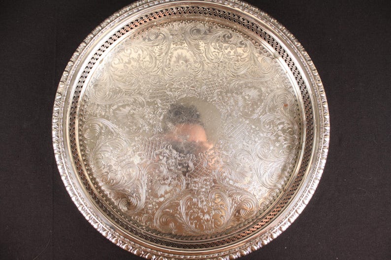 May include: A round, silver-colored serving tray with an ornate, embossed floral design. The tray has a decorative pierced border and a reflective surface. The tray is likely made of metal and is suitable for serving food or displaying items.