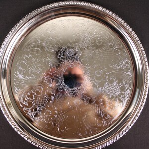 May include: A round, silver-colored serving tray with an ornate floral and scroll design. The tray has a decorative rope-like edge. The surface reflects the surrounding environment. The tray is likely made of metal.