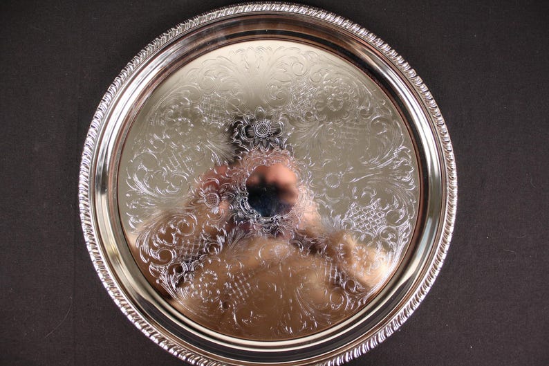 May include: A round, silver-colored serving tray with an ornate floral and scroll design. The tray has a decorative rope-like edge. The surface reflects the surrounding environment. The tray is likely made of metal.