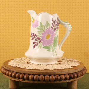May include: A cream-colored ceramic pitcher featuring a hand-painted floral design with purple and pink daisy-like flowers, green leaves, and dark purple sprigs. The pitcher has a scalloped rim, a curved handle, and a flared base.