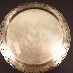May include: A silver-colored, round tray with a decorative, scalloped edge. The tray has a smooth, reflective surface and a detailed, openwork design along the rim. The tray is likely made of metal and is suitable for serving or display.
