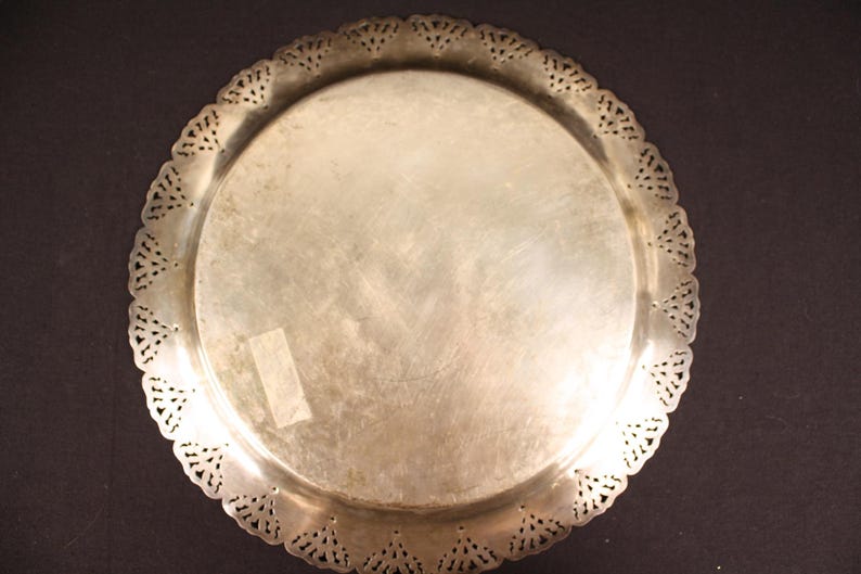 May include: A silver-colored, round tray with a decorative, scalloped edge. The tray has a smooth, reflective surface and a detailed, openwork design along the rim. The tray is likely made of metal and is suitable for serving or display.