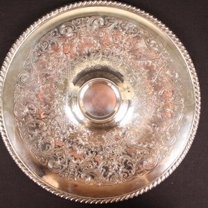 May include: A round, silver-colored serving tray with ornate floral and scrollwork patterns. The tray has a raised, decorative edge with a rope-like design. A smaller, circular indentation is in the center. The tray is likely made of metal.