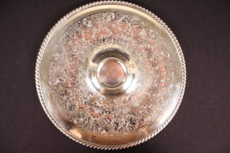 May include: A round, silver-colored serving tray with ornate floral and scrollwork patterns. The tray has a raised, decorative edge with a rope-like design. A smaller, circular indentation is in the center. The tray is likely made of metal.