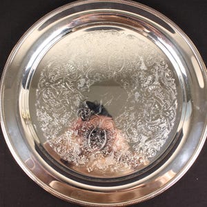 May include: A round, silver-colored serving tray with an ornate floral pattern in the center. The tray has a beaded edge and a reflective surface. The tray is likely made of metal and is suitable for serving food or displaying items.