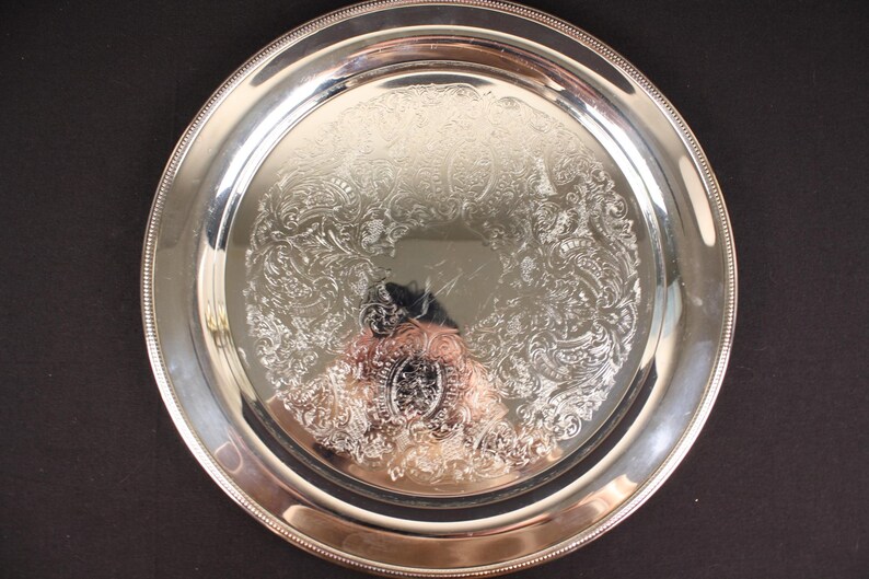 May include: A round, silver-colored serving tray with an ornate floral pattern in the center. The tray has a beaded edge and a reflective surface. The tray is likely made of metal and is suitable for serving food or displaying items.