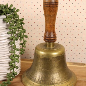 May include: A vintage brass bell with a wooden handle. The bell has a tarnished, aged appearance and the word "UNCLEAN" is engraved on the base. The handle is a dark brown color. The bell is likely used for decorative purposes.