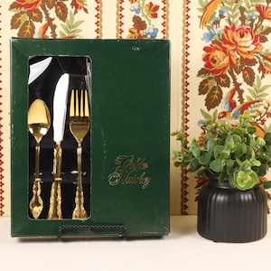 May include: A boxed set of gold-colored flatware, including a spoon, knife, and fork, is displayed in a dark green box. The box has a clear window. The words "Gold Matchy" are printed on the box. A small black vase with greenery is to the right.