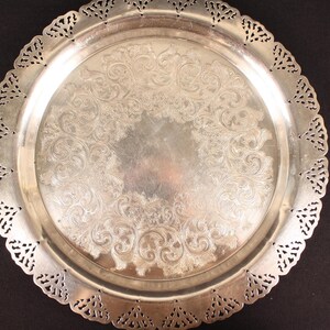 May include: A silver-colored, round serving tray with an ornate, embossed floral design in the center. The tray has a decorative, openwork edge. The tray is likely made of metal and is suitable for serving food or displaying decorative items.
