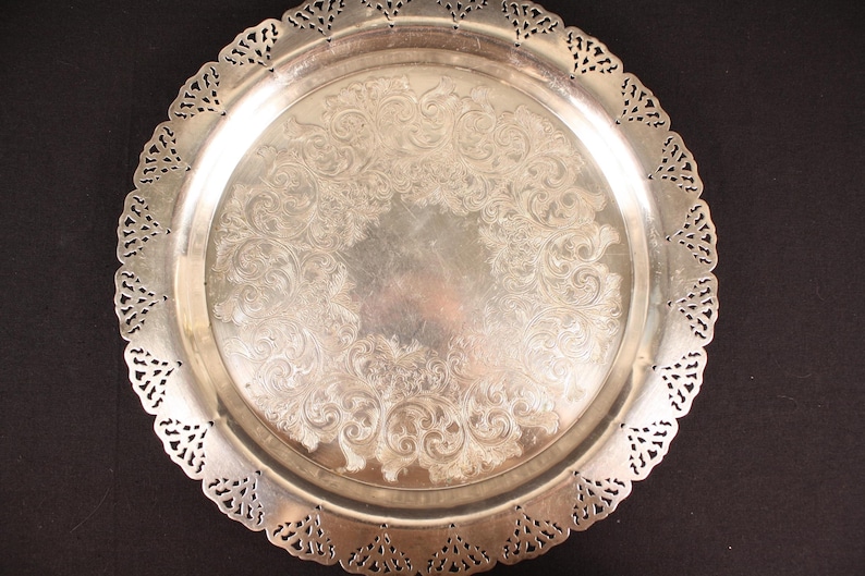 May include: A silver-colored, round serving tray with an ornate, embossed floral design in the center. The tray has a decorative, openwork edge. The tray is likely made of metal and is suitable for serving food or displaying decorative items.