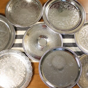 May include: An assortment of silver-colored serving trays with ornate, embossed designs. The trays vary in size and shape, some with beaded edges and others with scalloped rims. They are arranged on a striped runner and wooden surface.