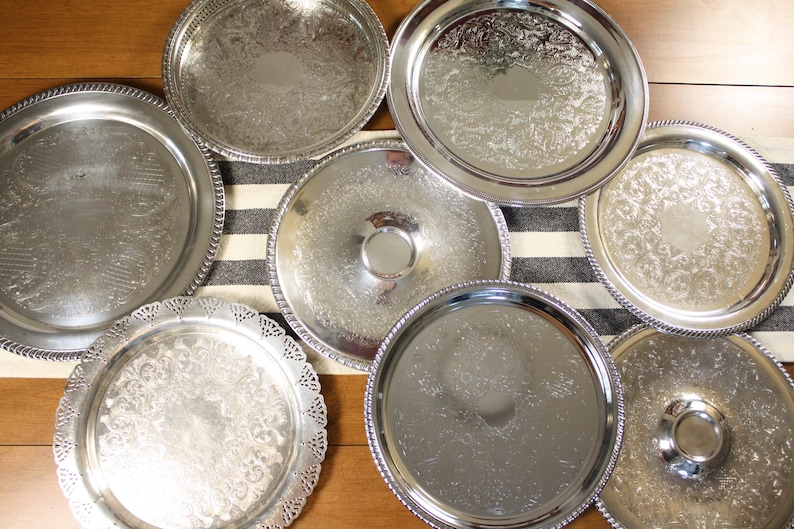 May include: An assortment of silver-colored serving trays with ornate, embossed designs. The trays vary in size and shape, some with beaded edges and others with scalloped rims. They are arranged on a striped runner and wooden surface.