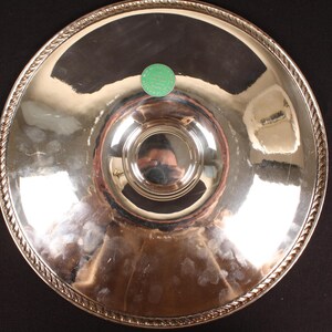 May include: A silver-colored serving tray with a decorative rope-like border. The tray has a central, raised circular area and a small green sticker. The reflective surface shows distorted reflections.