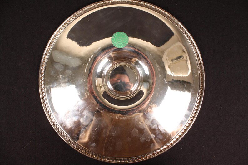 May include: A silver-colored serving tray with a decorative rope-like border. The tray has a central, raised circular area and a small green sticker. The reflective surface shows distorted reflections.