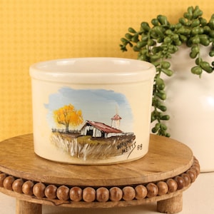 May include: A cylindrical cream-colored ceramic crock featuring a hand-painted landscape scene of a barn, a silo, and a yellow tree. The artwork includes the signature Wayne Metts and the year 88. The vessel has a smooth, matte finish.