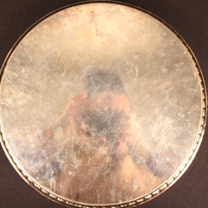 May include: A round, silver-toned metal tray with a decorative beaded edge. The tray's surface is reflective, showing a distorted reflection. The tray is set against a dark background.