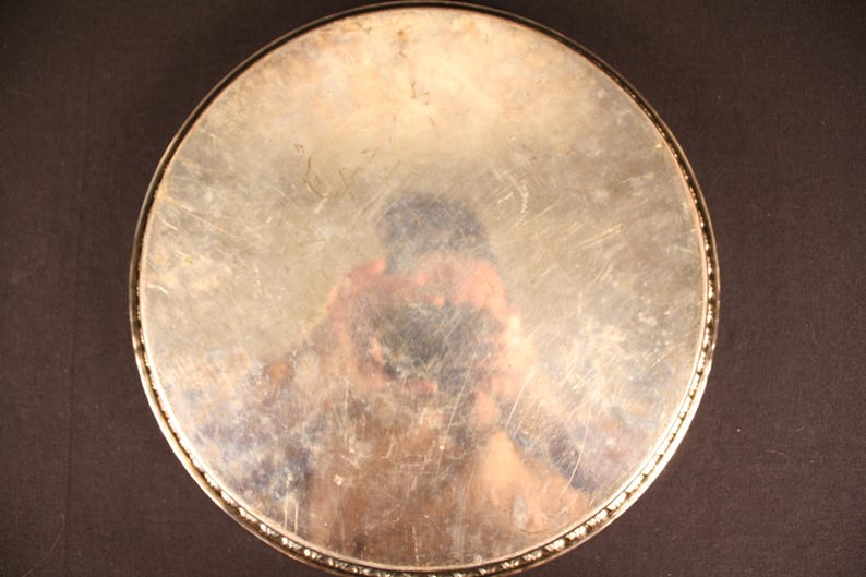 May include: A round, silver-toned metal tray with a decorative beaded edge. The tray's surface is reflective, showing a distorted reflection. The tray is set against a dark background.