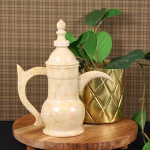 May include: A beige marble-like coffee pot with a curved handle and a decorative top. It sits on a wooden stand next to a gold-colored, diamond-patterned pot holding a green plant. The background is a brown plaid pattern.