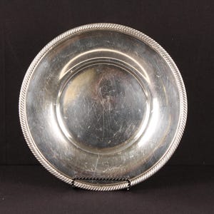May include: A round silver-toned metal bowl featuring a polished, reflective interior surface with visible surface scratches and a decorative rope-style texture along the outer rim. The bowl has a smooth, concave center and a wide, flat edge.