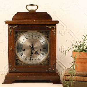 May include: A vintage wooden mantel clock with a silver face and Roman numeral hour markers. The clock has a dark brown finish, decorative metal corner accents, and a handle on top. A small potted plant sits next to the clock.