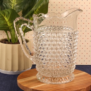 May include: A clear glass pitcher with a hobnail design, featuring a handle and a spout. The pitcher sits on a wooden round board. A potted plant with green leaves is in the background.