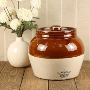 May include: A two-toned ceramic crock with a glossy brown glazed upper section and a matte cream-colored lower section. The vessel features a matching lid and a small blue stamped crown logo with the number three on the front lower exterior.