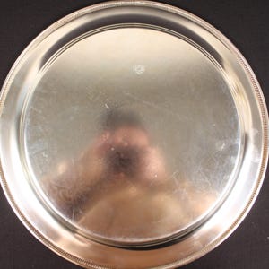 May include: A round, silver-colored serving tray with a beaded edge. The tray has a reflective surface and a slightly raised rim. The background is a solid black color.