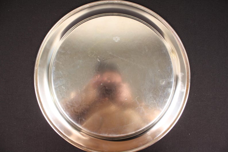 May include: A round, silver-colored serving tray with a beaded edge. The tray has a reflective surface and a slightly raised rim. The background is a solid black color.