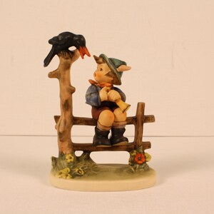May include: A ceramic figurine depicting a young boy playing a horn while seated on a wooden fence. A black bird perches on a tree branch above him. The figurine has a detailed, hand-painted design with a base of flowers.