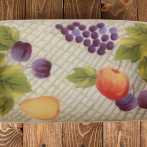 May include: A rectangular ceramic serving platter featuring a hand-painted fruit motif. The design includes clusters of purple grapes, a yellow pear, a red apple, and several purple plums surrounded by green leaves against a light lattice background.