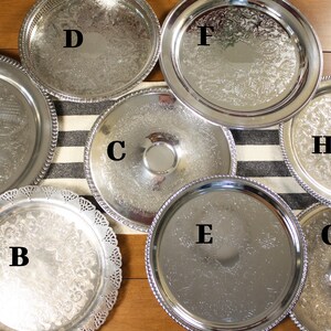 May include: An assortment of silver serving trays with various designs and edges. The trays are round and feature ornate patterns, beaded edges, and a central indentation. The trays are arranged on a striped runner and wooden surface.