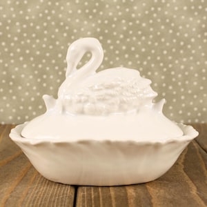May include: A white ceramic lidded dish featuring a sculpted swan figure on the top. The lid has a smooth finish and sits atop a bowl with a scalloped edge. The swan is depicted with its neck curved and detailed feathers on its body.