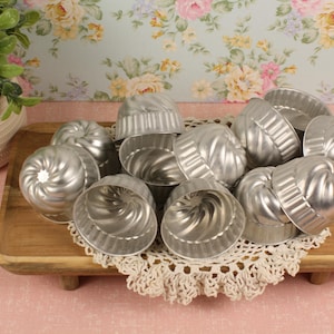 May include: A collection of small, silver-colored metal bundt cake molds. The molds are arranged on a wooden tray atop a cream-colored lace doily. A potted plant is visible in the background, with a floral wallpaper.